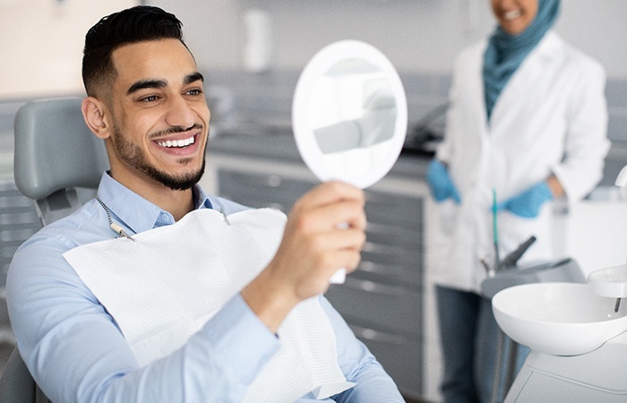 Man smiling at reflection in handheld mirror with dentist in background