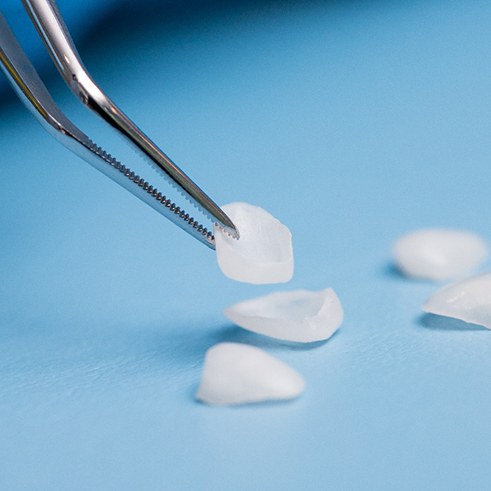 Tweezers picking up a veneer from a pile of several on a pale blue surface